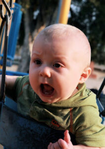 A baby in a park swing, mouth wide open like he's screaming