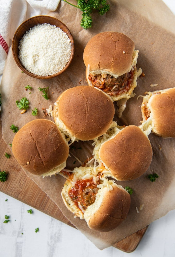 an overhead image with 6 meatball sliders on brown paper and a wooden cutting board wit a small bowl of grated parmesan.