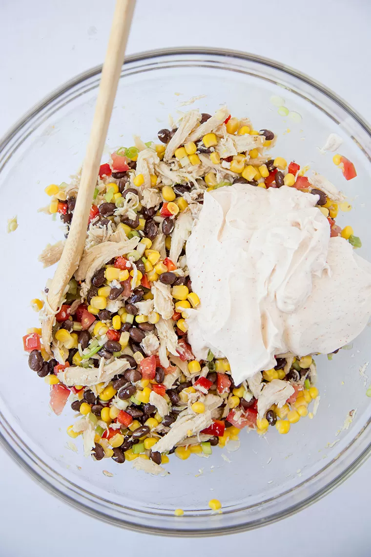Skinny Southwest Chicken Dip is full of shredded chicken and loads of veggies and spices for a healthy Southwest Chicken Salad appetizer or main course! southwest chicken dip ingredients in a bowl with a wooden spoon.