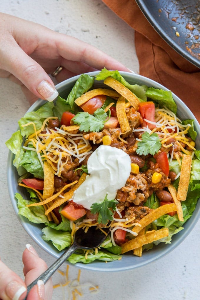 A bowl filled with salad and taco meat. Two hands, one holding the bowl and the other holding a fork with a bite of salad on it.