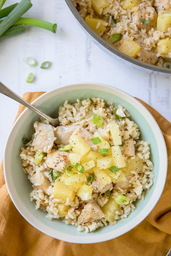 A blue bowl with chicken, pineapple and rice topped with sliced green onions. A fork sticks out of the bowl.