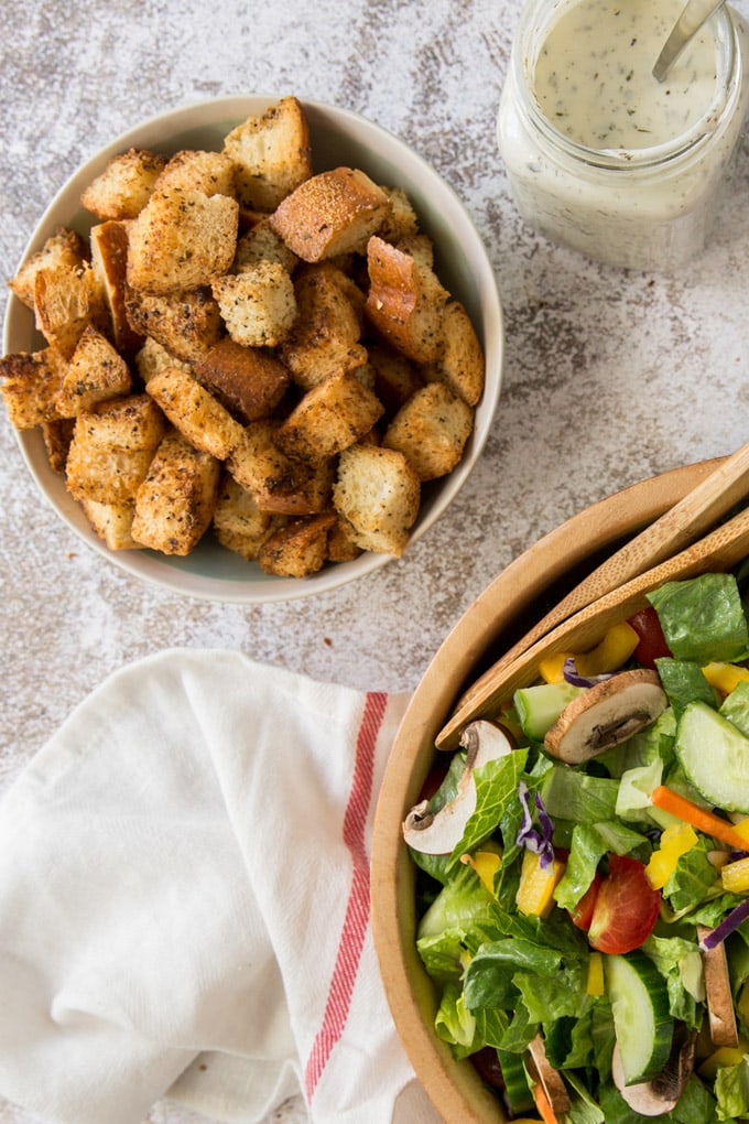 a bowl of croutons, a wood bowl of salad, a jar of ranch dressing, a white and red towel