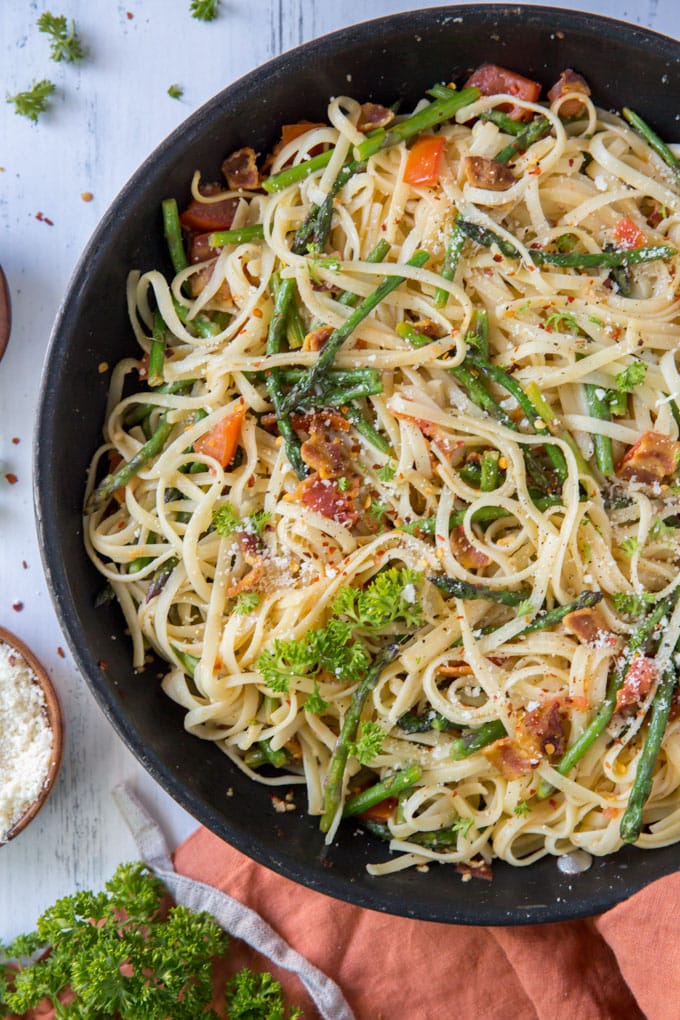 Linguine pasta in a skillet with bacon, asparagus, tomatoes and red pepper flakes sitting on an orange napkin.