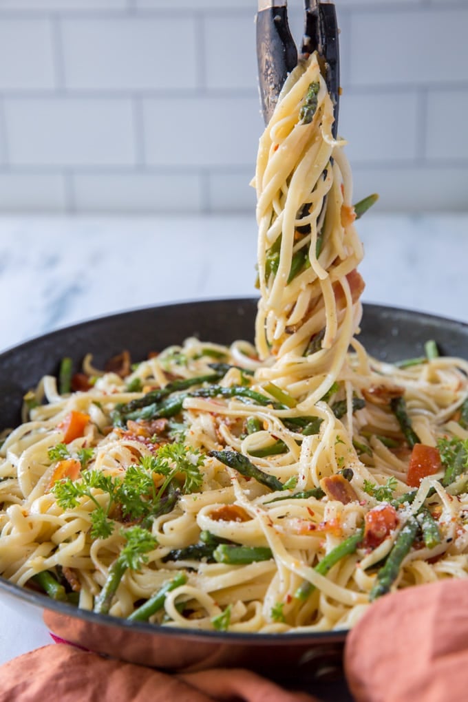 Linguine pasta in a skillet with bacon, asparagus, tomatoes and red pepper flakes sitting on an orange napkin. Tongs lift some pasta out of the pan.