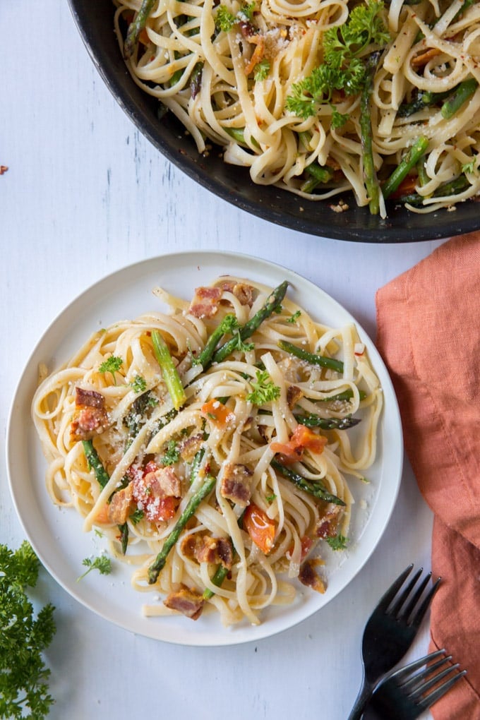 a white dinner plate holding linguine pasta with bacon, asparagus, tomatoes and Parmesan cheese sitting next to an orange napkin and a skillet holding the same pasta.