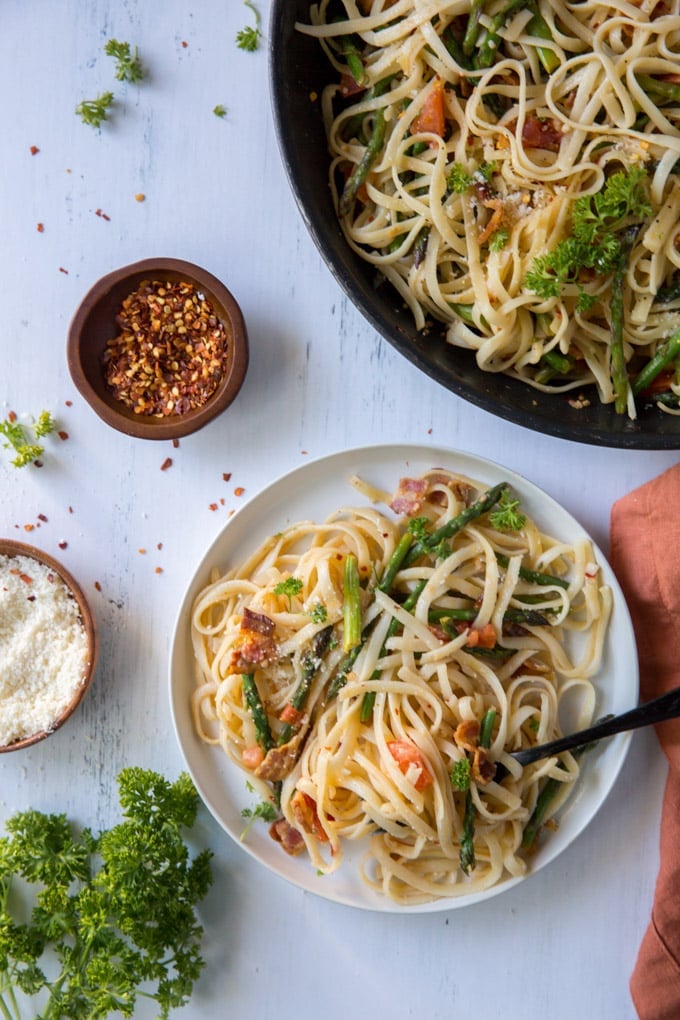 a white dinner plate with a fork twirling linguine pasta with bacon, asparagus, tomatoes and Parmesan cheese sitting next to an orange napkin and a skillet holding the same pasta. There is also a small bowl with red pepper flakes, and another with parmesan cheese.