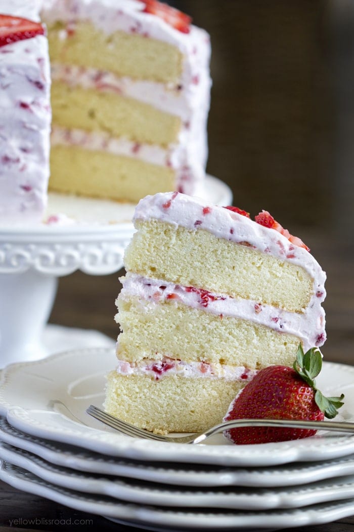 Fresh Strawberry Cake - a dense and moist vanilla cake with three layers, and a light and fluffy whipped cream strawberry frosting. a slice of vanilla cak.e with straberry frosting sitting on a plate with a strawberry and a fork