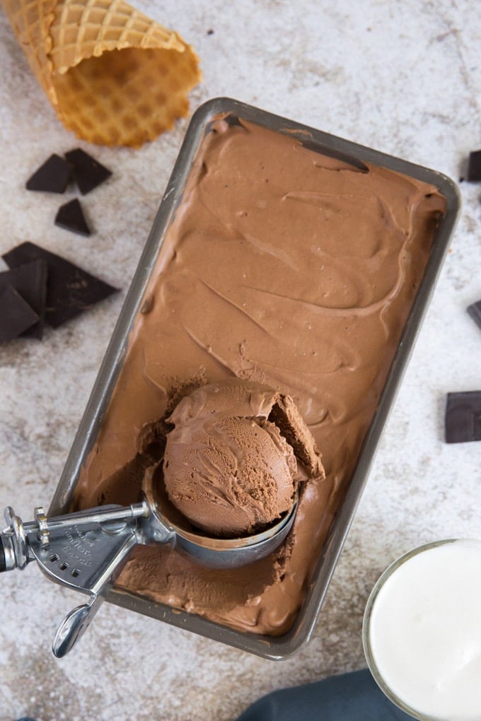 a loaf pan with chocolate ice cream, an ice cream scoop, broken pieces of chocolate, a waffle cone.