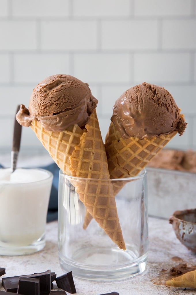 a glass with two ice cream cones topped with chocolate ice cream. a jar of marshmallow sauce in the background. Pieces of chopped chocolate