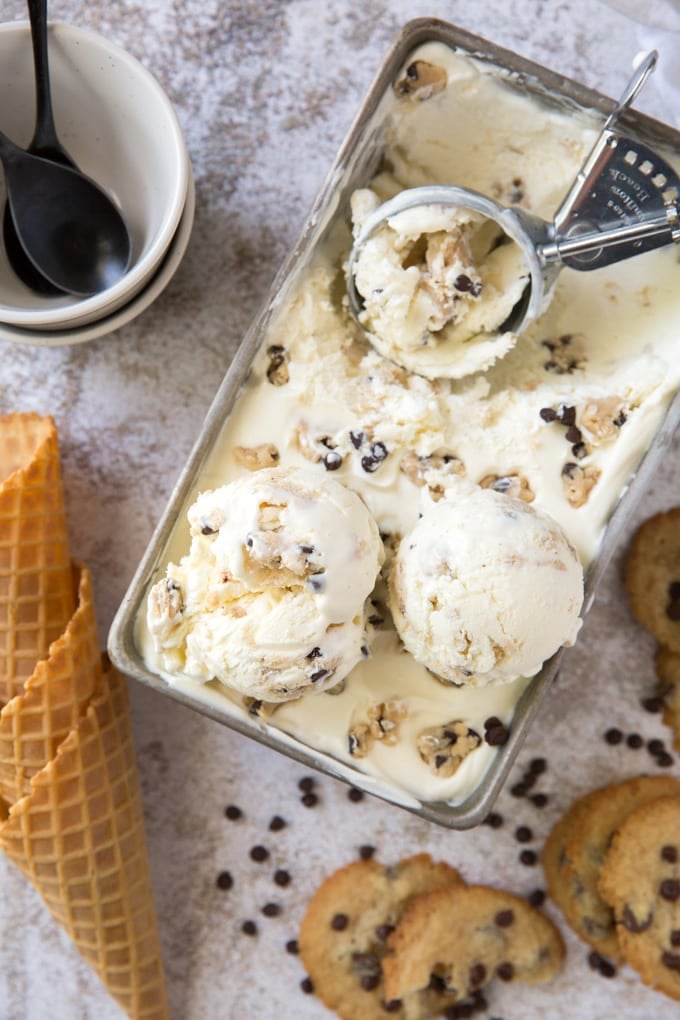 An overhead image with 2 stacked white bowls with a black spoon, 3 ice cream cones, scattered chocolate chips and chocolate chip cookies and a loaf pan with cookie dough ice cream and an ice cream scoop.