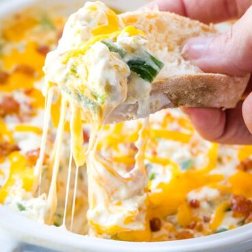 hand holding piece of bread with jalapeno, cheese pull, dip in white dish
