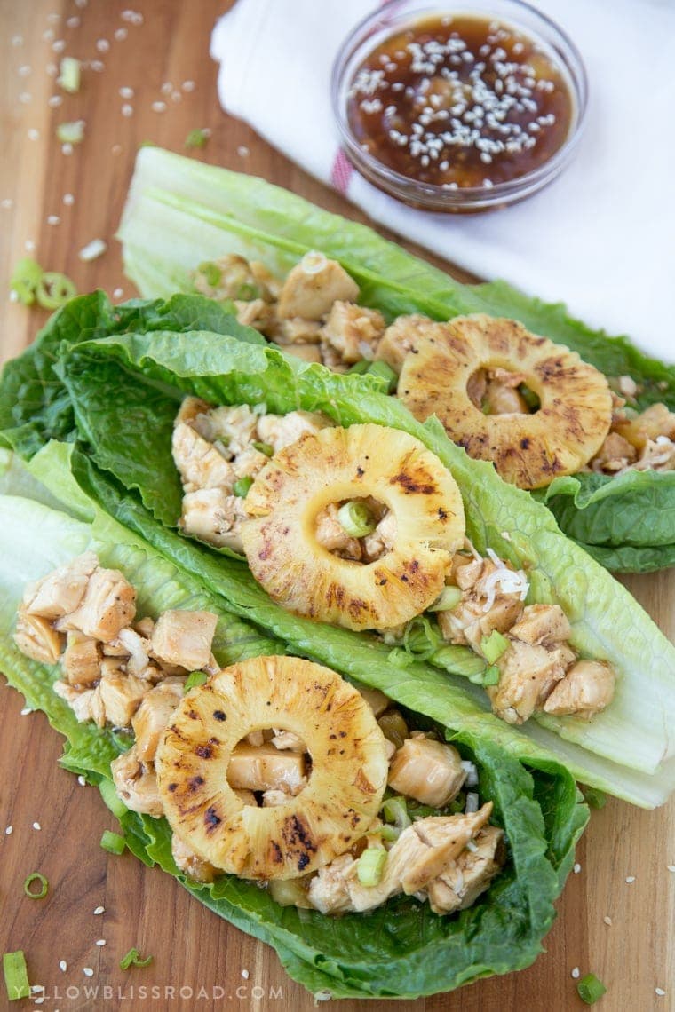 teriyaki sauce in a small dish with sesame seeds on top, a wood cutting board, 3 romaine lettuce leaves with chicken and a slice of pineapple in each.