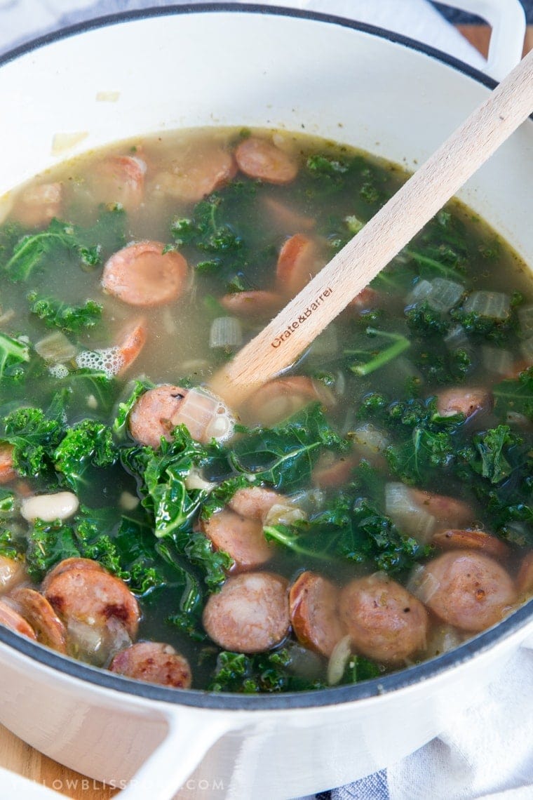 Andouille sausage and kale soup in a large white pot with a wooden spoon