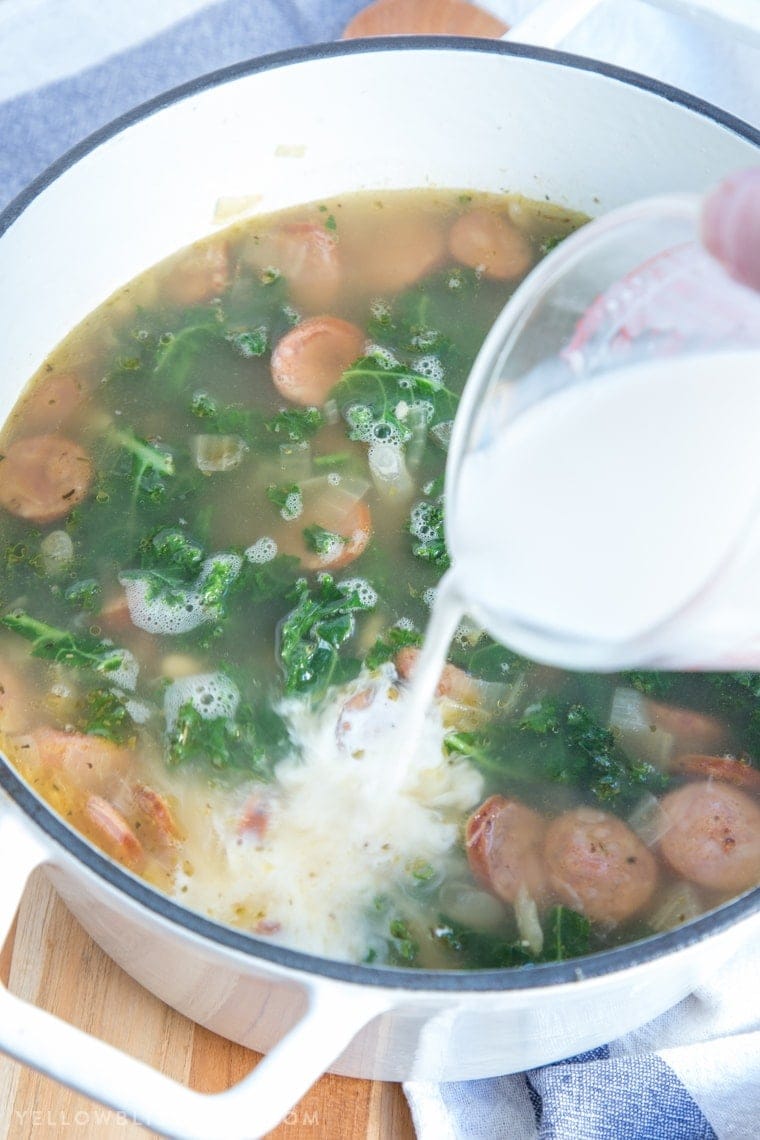 Half and half being poured into a pot filled with sausage, kale, beans and chicken broth