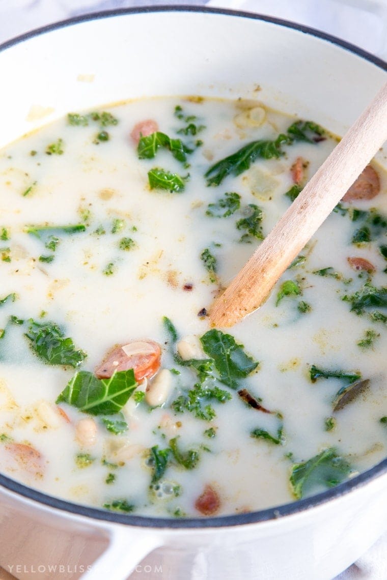 A pot filled with creamy broth, white beans, andouille sausage and kale with a wooden spoon