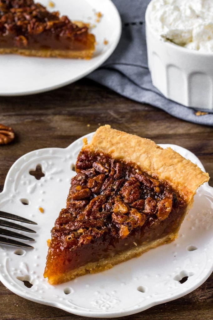 This classic Pecan Pie Recipe is filled with crunchy pecans baked with a sweet filling. This beautiful pie will steal the show on your dessert table. A slice of pecan pie on a white plate.
