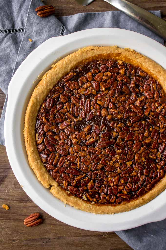 This classic Pecan Pie Recipe is filled with crunchy pecans baked with a sweet filling. This beautiful pie will steal the show on your dessert table. Pecan Pie recipe in a white pie plate.