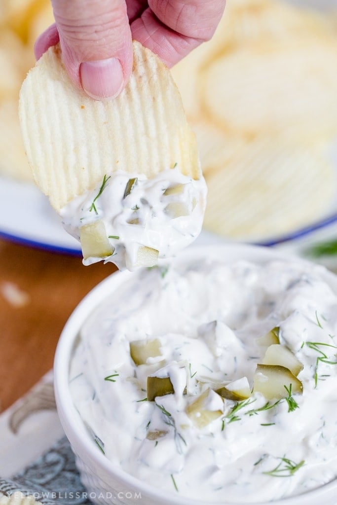 A hand reaching in holding a potato chip with dill pickle dip on it.