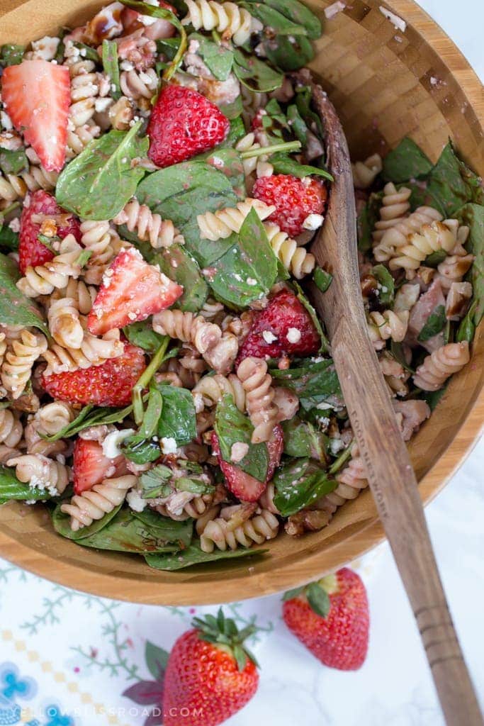 a large wood bowl with rotini pasta, strawberries, spinach nuts and cheese.