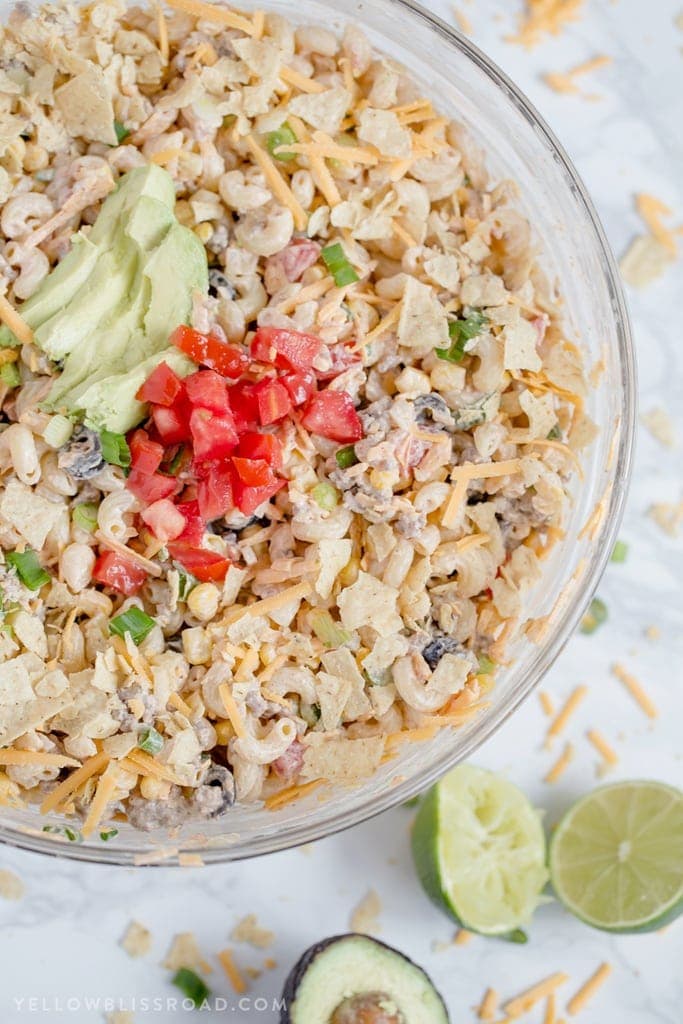 a large clear glass bowl with macaroni salad with black beans, taco meat, tomatoes, avocado and limes.