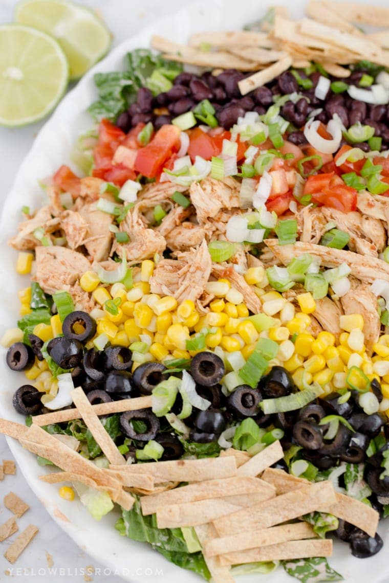 a white platter with lettuce, tortilla strips, olives, corn, chicken, tomatoes and black beans
