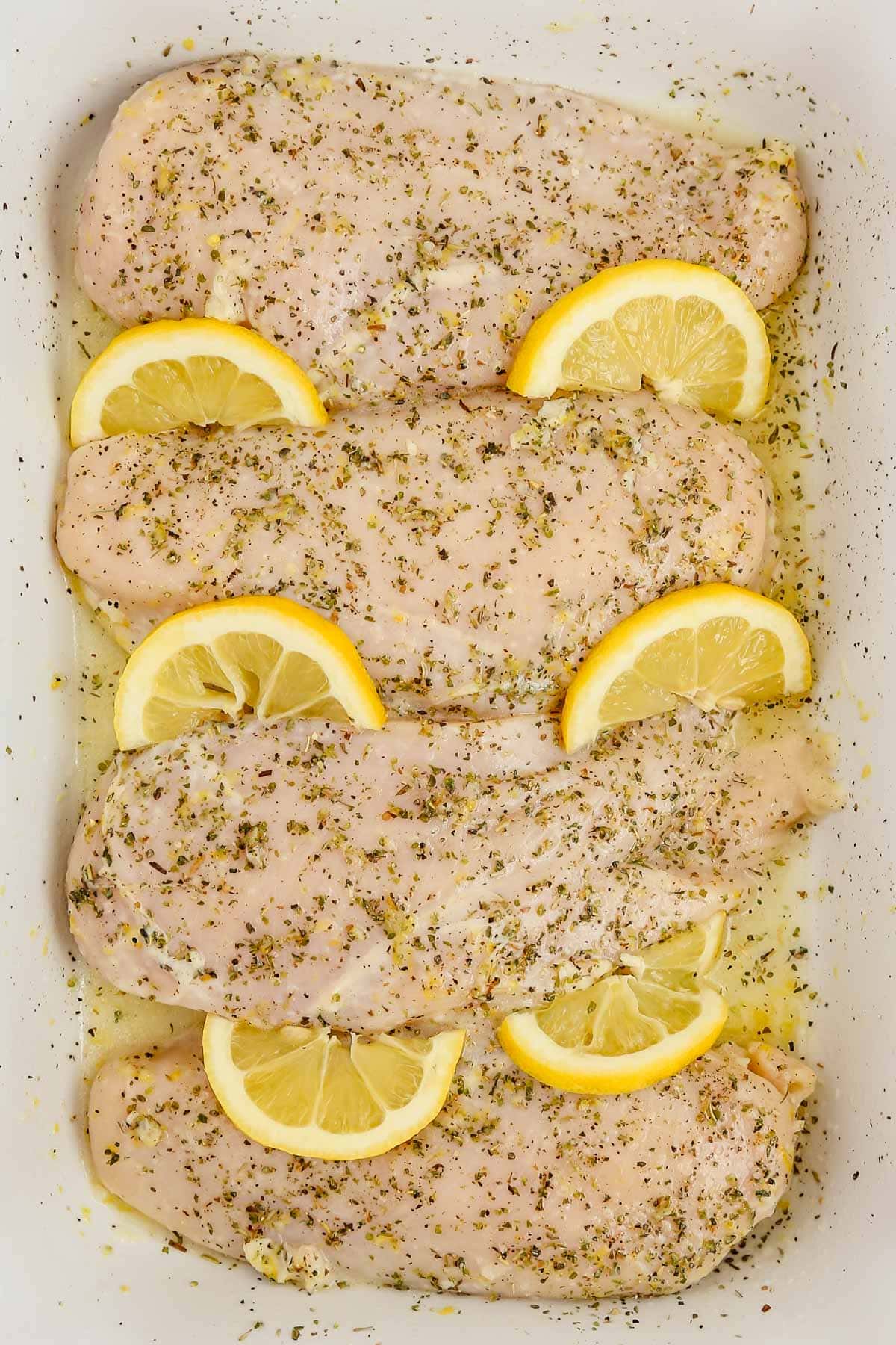 4 uncooked chicken breasts with lemon slices and seasoning.