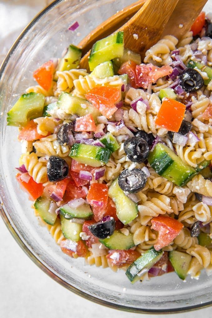 An overhead shot of greek pasta salad with salad tongs sticking out of the bowl