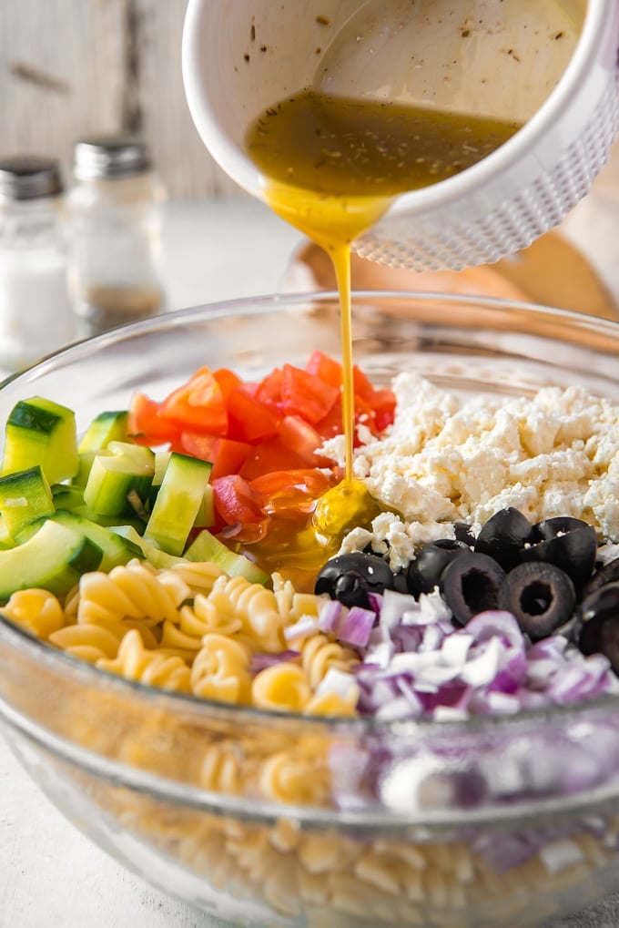 Ingredients to make greek pasta salad in a bowl with dressing being poured on.