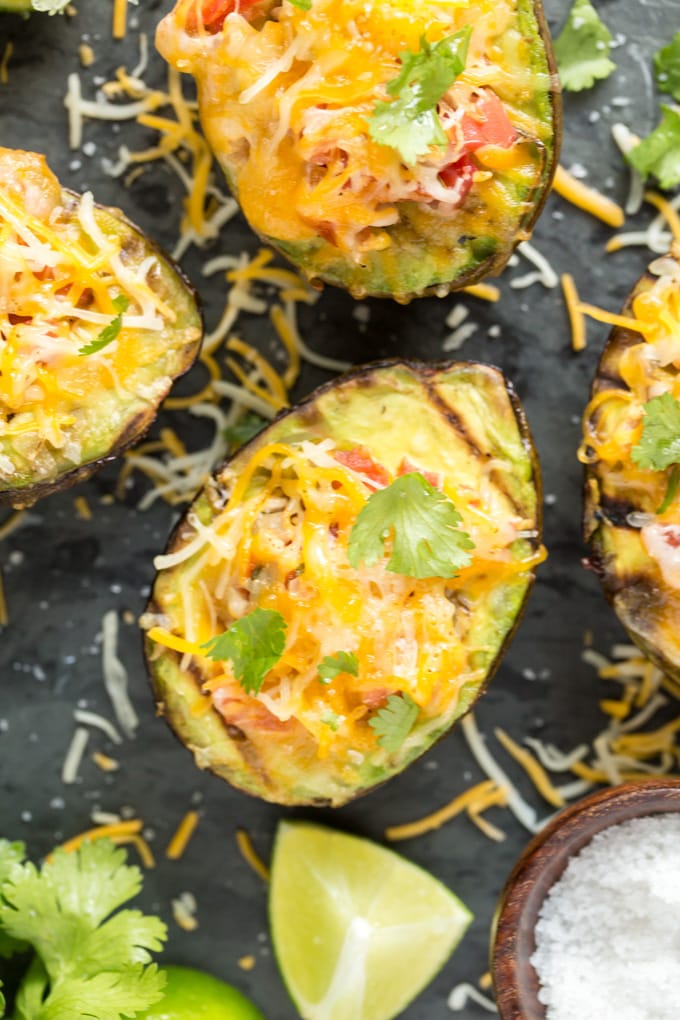 Grilled avocados on a black background topped with cheese, salsa and cilantro. A small bowl of coarse salt and some sliced limes surround them.