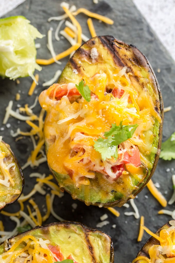 Grilled avocado halves filled with salsa and cheese and topped with cilantro leaves, sitting on a black cutting board.