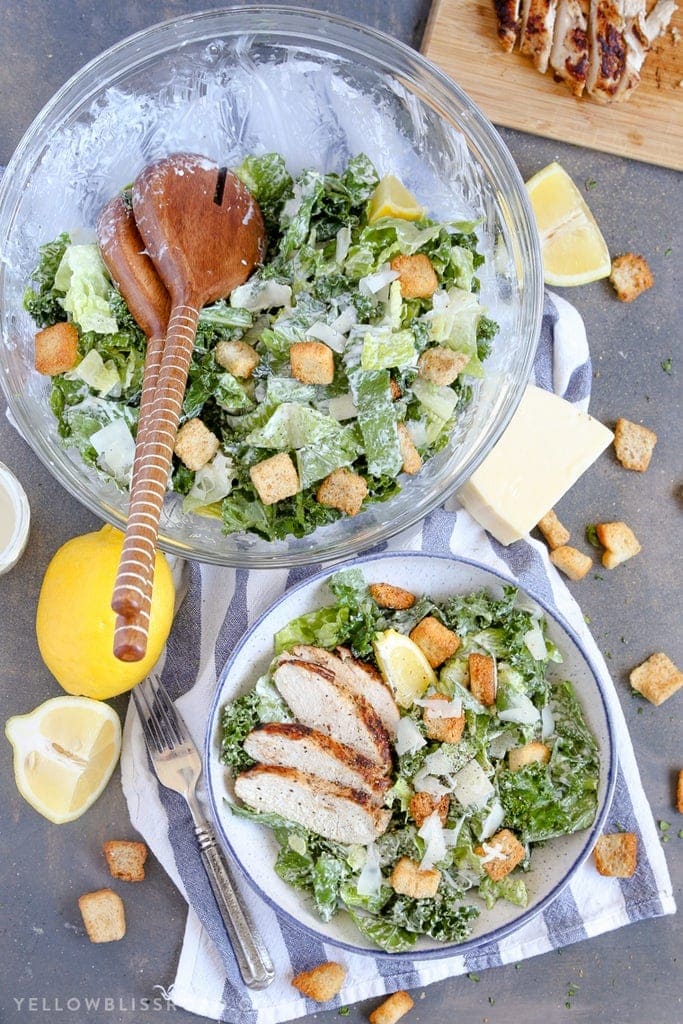 a large bowl of caesar salad, a small bowl ow casear salad with slices of chicken, a blue and white napkin and lemon slices.