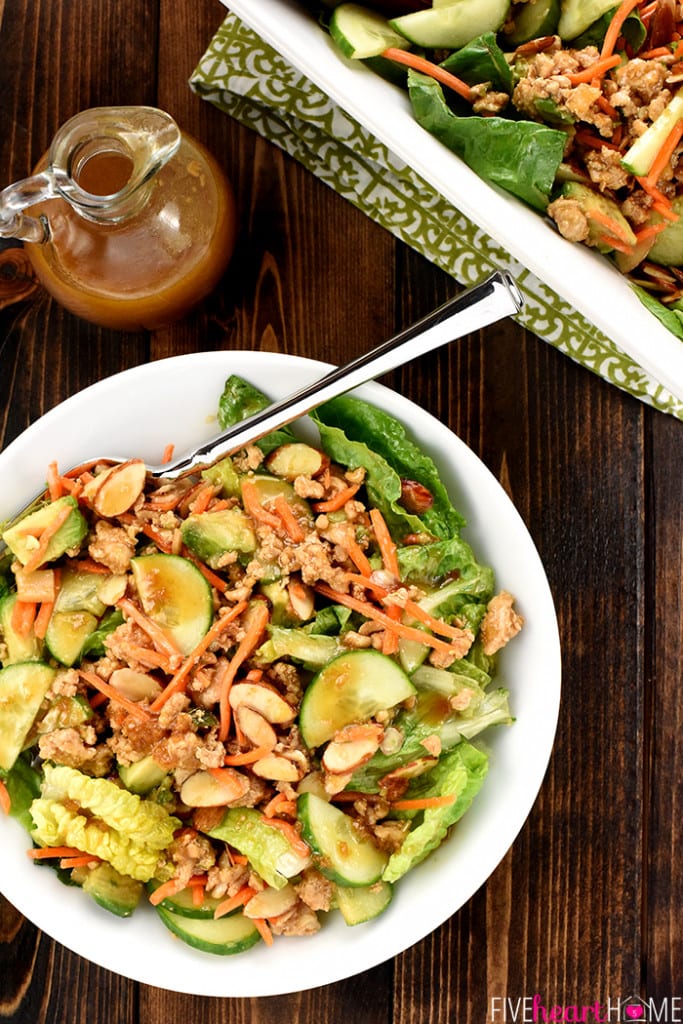 a white bowl with ground chicken, cucumbers, lettuce and carrots and a spoon. A small jar of dressing. A wood background