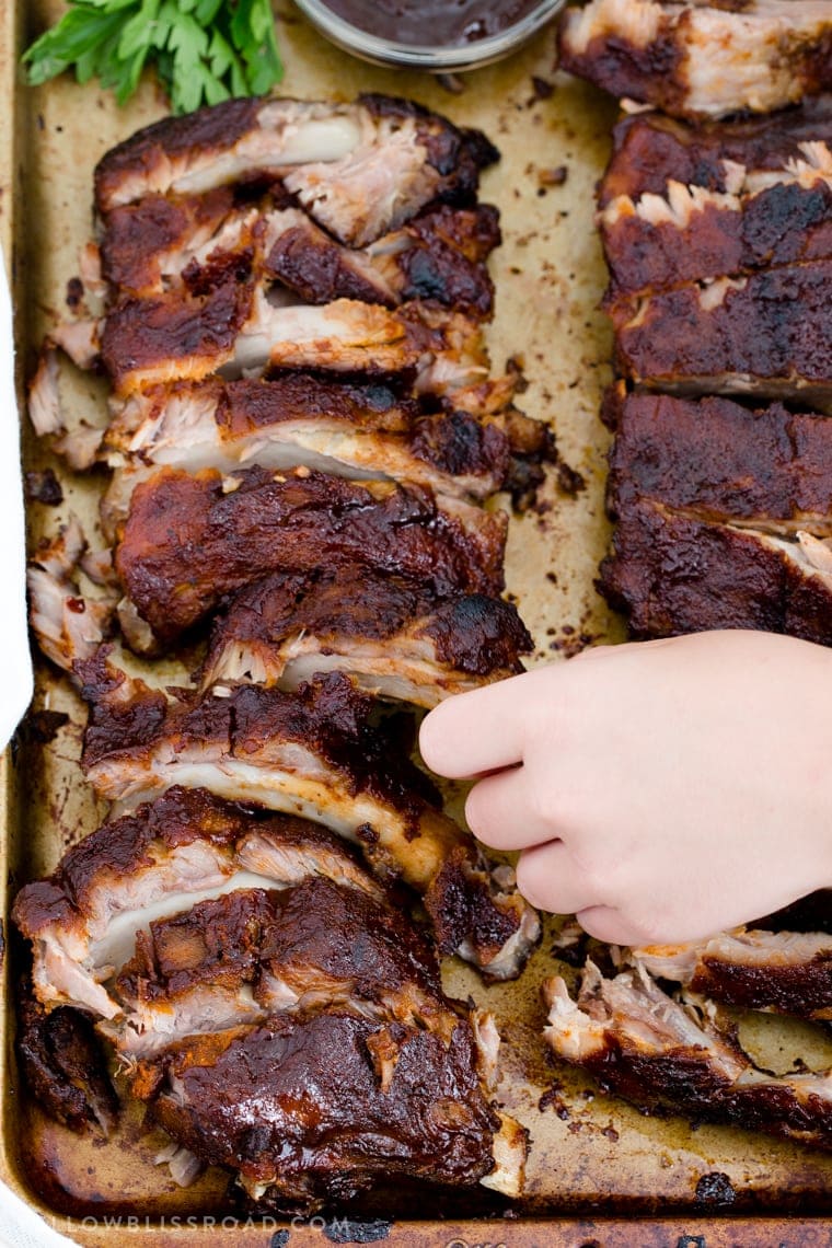 slow cooker ribs with barbecue sauce on a baking sheet. A little hand is trying to grab a rib.
