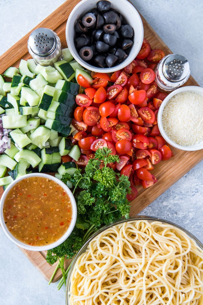 a wood cutting board, a gray background, a bowl of cooked spaghetti noodles, small bowls of Italian dressing, parmesan cheese and olives, chopped tomatoes and cucumbers, parsley and salt and pepper shakers.