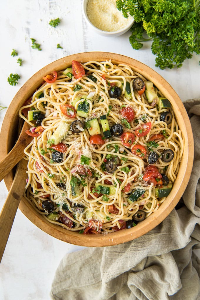 a tan napkin, parsley, a bowl of parmesan cheese, a wooden bowl with wooden spoons, spaghetti, cucumbers, olives and tomatoes.