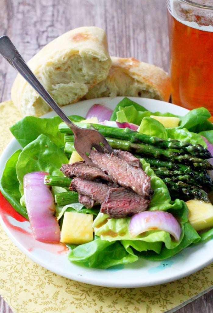 a small white plate with lettuce, onions, asparagus and sliced steak. A glass with tea and a piece of bread.