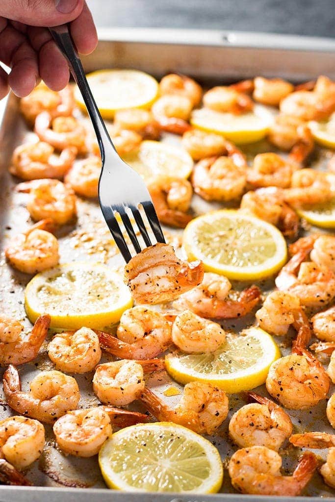 A fork holding two shrimp over a sheet pan full of lemon pepper shrimp