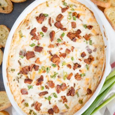 Creamy bacon dip with bacon crumbles, green onions in a white baking dish, crostini.