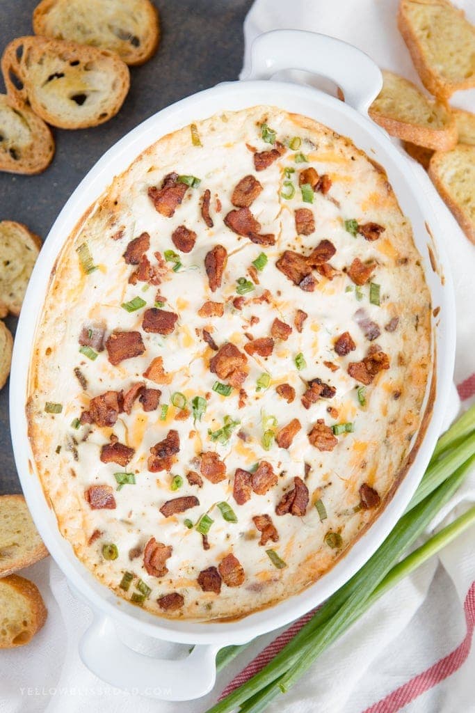 Creamy bacon dip with bacon crumbles, green onions in a white baking dish, crostini. 