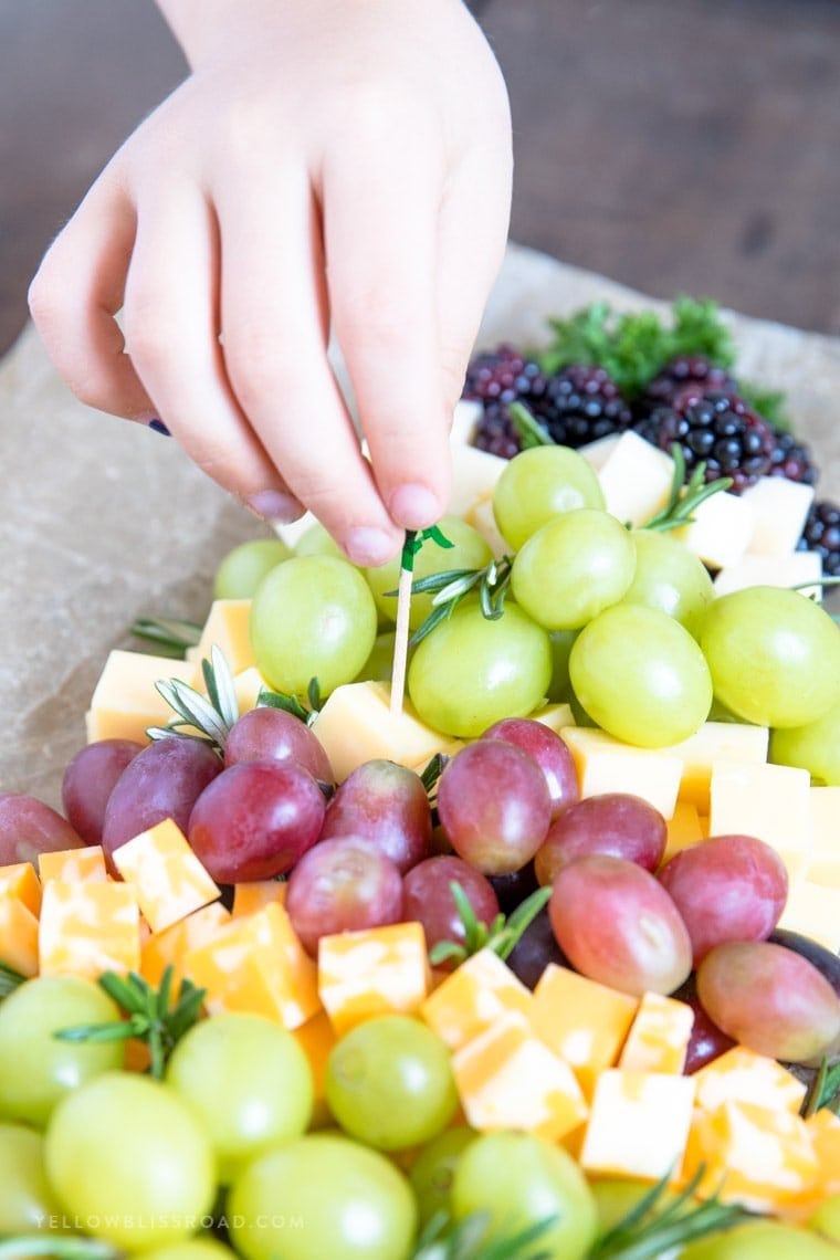 A child's hand reaching in to grab a piece of cheese from a fruit and cheese platter