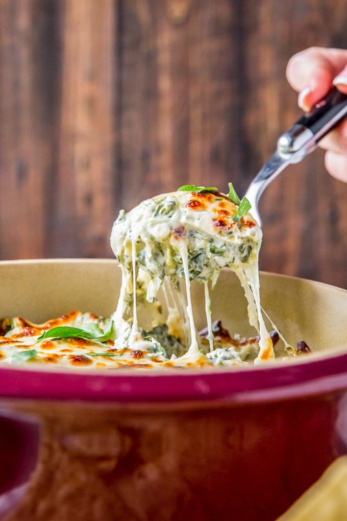 A spoon lifting a serving of spinach artichoke dip from a baking dish.