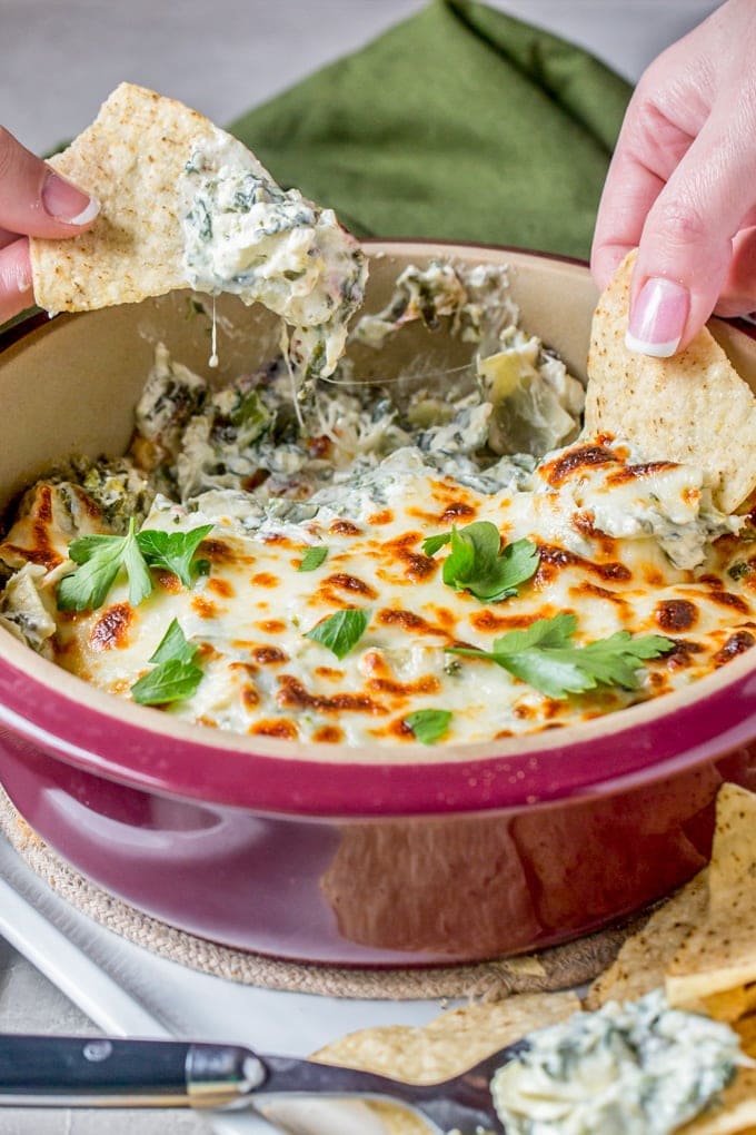 Two hands dipping tortilla chips into a pan of spinach artichoke dip.