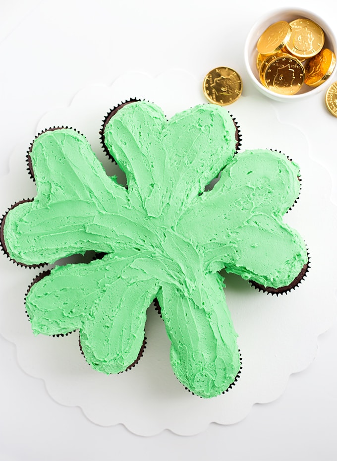 This sweet Shamrock Cake is the perfect St. Patrick's Day Dessert! A simple pull-apart cake with chocolate cupcakes, green frosting and rainbow sprinkles. Green frosted cupcakes on a white cake board in the shape of a shamrock
