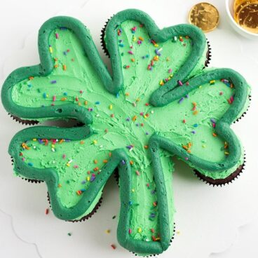 Green frosted cupcakes on a white cake board in the shape of a shamrock with a dark green outlining the shamrock and rainbow sprinkles on top