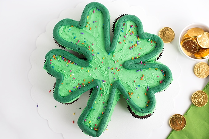 Green frosted cupcakes on a white cake board in the shape of a shamrock with a dark green outlining the shamrock and rainbow sprinkles on top