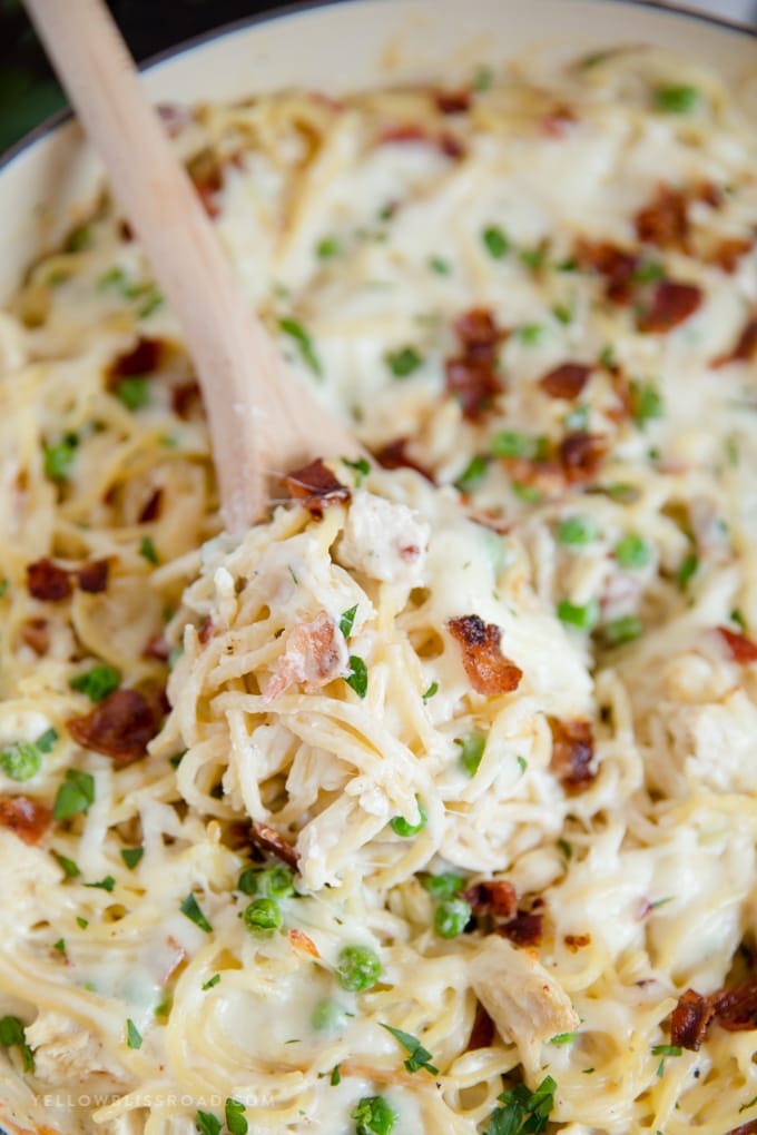 Creamy, Cheesy Chicken Spaghetti, with bacon, chicken, spaghetti and peas in a made from scratch sauce. Pure comfort food and perfect for a hearty weeknight meal. A wooden spoon in a pan of cheesy chicken spaghetti with bacon.