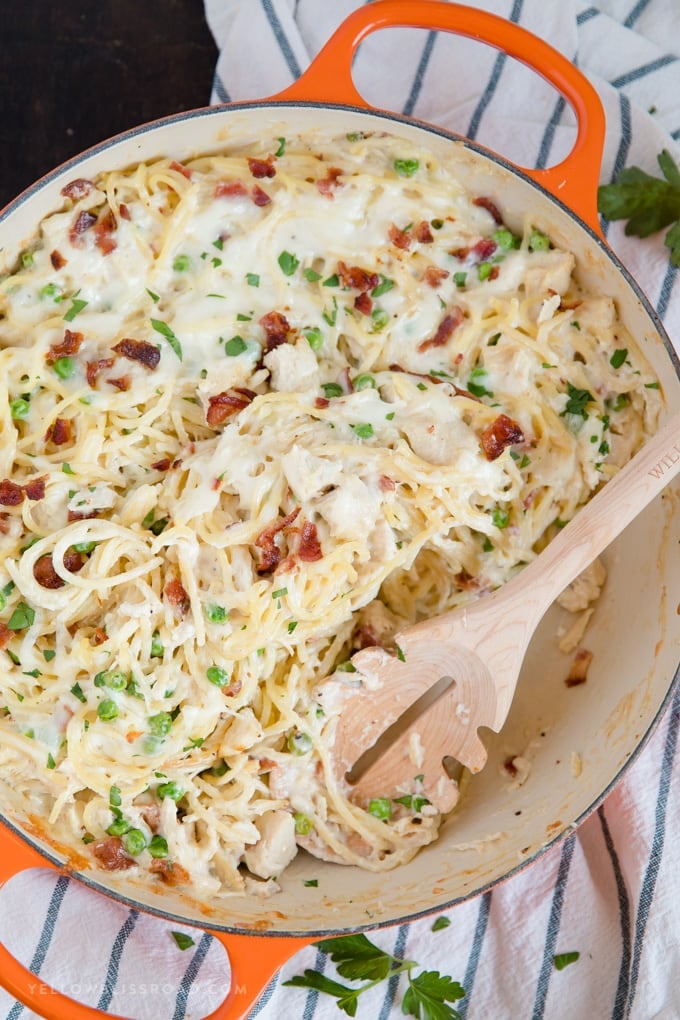 A pan of Creamy, Cheesy Chicken Spaghetti, with bacon, chicken, spaghetti and peas in a made from scratch sauce with a wooden spoon.