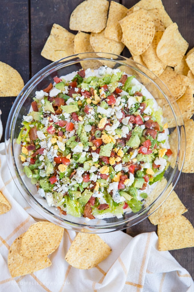 cobb salad dip in a clear dish surrounded by tortilla chips and a yellow and white towel