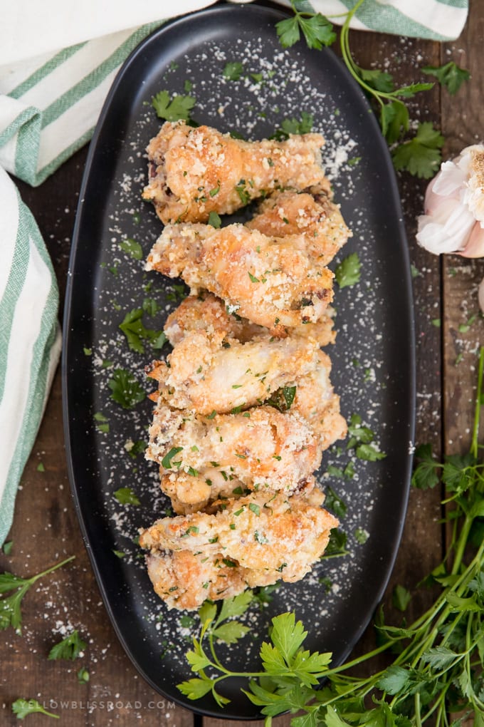 Crispy Baked Garlic Parmesan Chicken Wings in garlic parmesan sauce stacked on a black platter with parmesan and parsley on a wood background