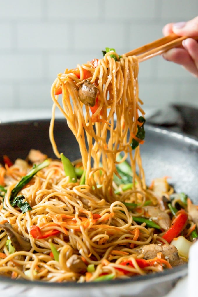 A hand holding chopsticks lifting up a serving of chicken lo mein from a large skillet